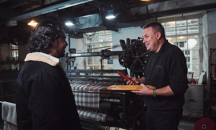 A craftsman explains the weaving process to a visitor. He holds a bobbin of red thread and a wooden shuttle, which passes through the loom to weave th